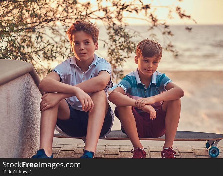 Portrait of two little brothers sitting together on a skateboard against the background of the seacoast at a sunset.