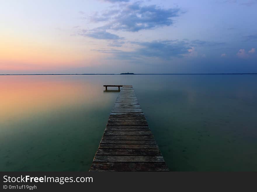 An old pier reveals views of the charmed lake, blue sky with cloud. Sunrise enlightens the horizon with orange warm colors.