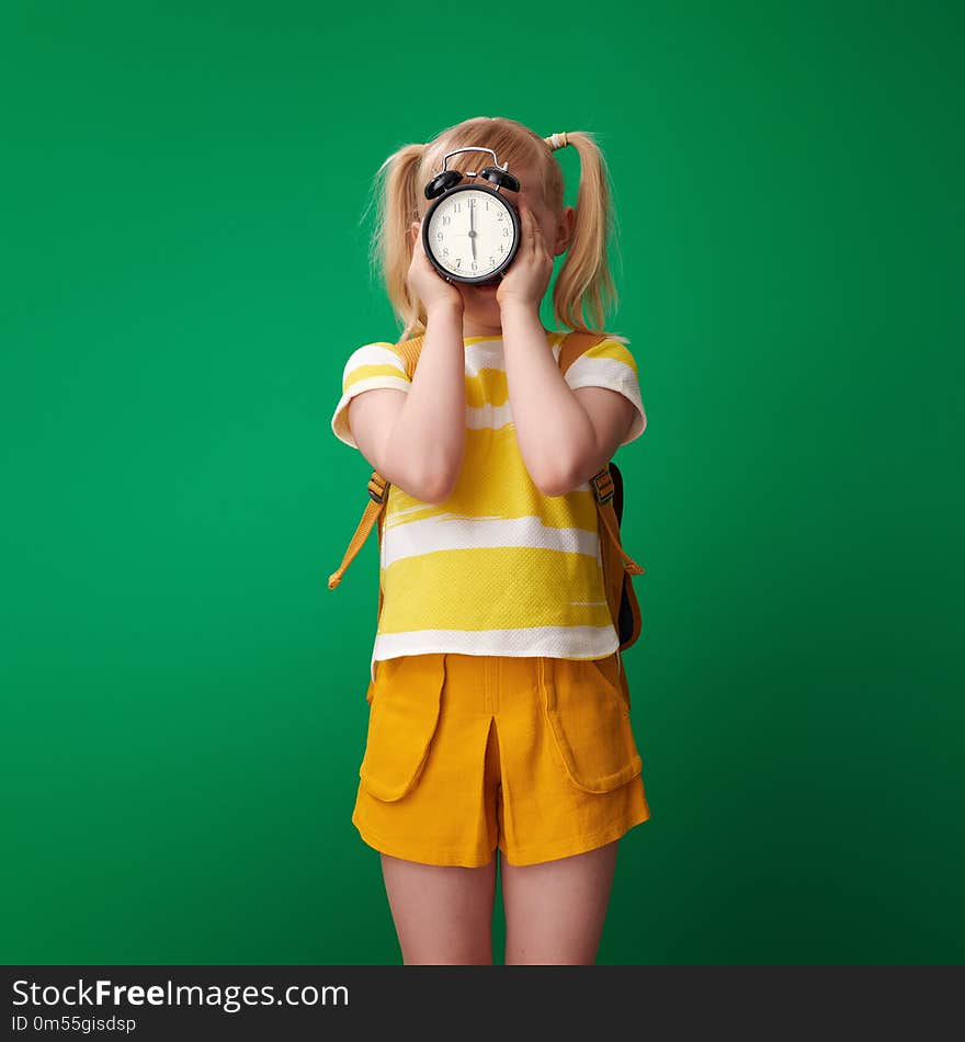 School girl with backpack holding alarm clock in the front of face against green background. School girl with backpack holding alarm clock in the front of face against green background