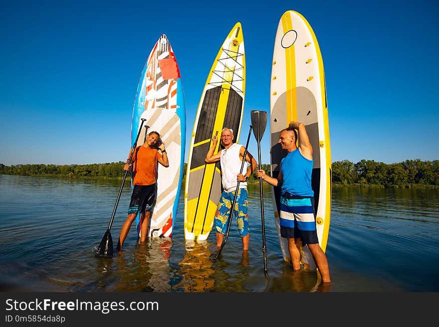 Joyful friends with SUP boards in their hands stand on the bank of a big river in the summer evening and enjoy life.