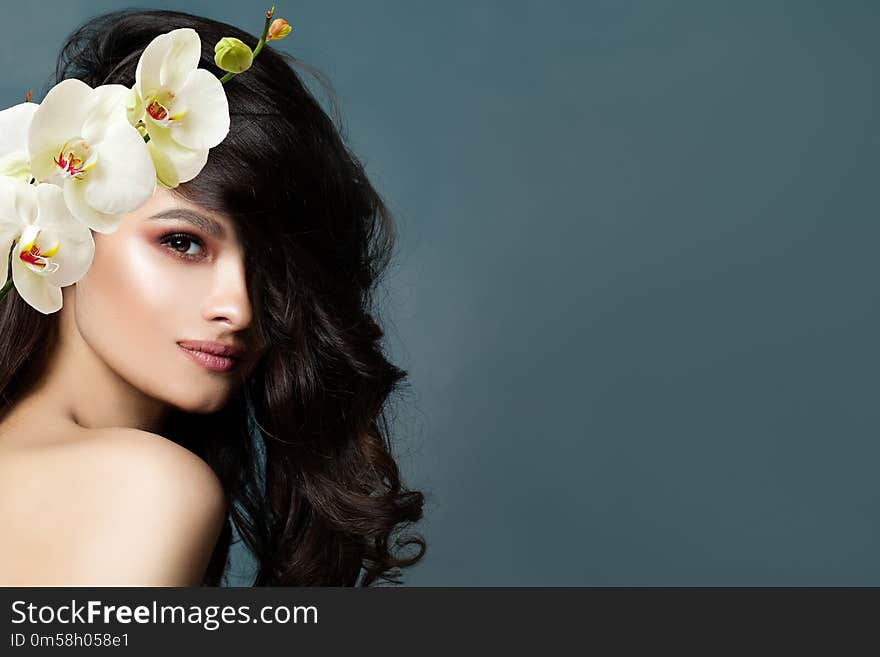 Beautiful brunette woman with healthy skin, dark wavy hair and white orchid flowers on blue background with copy space. Facial treatment, cosmetology, beauty salon, skin care and spa