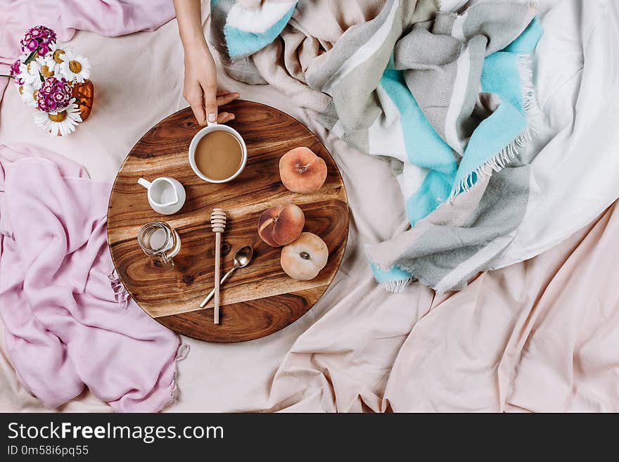 Cozy autumn or winter flatlay of wooden tray with cup of coffee, peaches, creamer with plant milk, syrup and spoons on pastel sheets and woman`s hand, selective focus