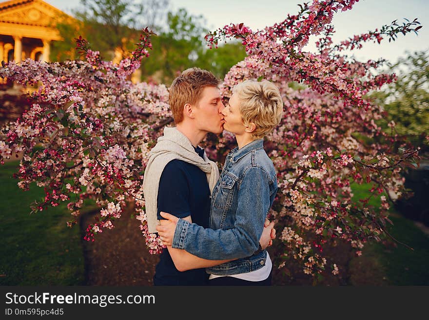 Stylish and beautiful women with short light hair, dressed in a blue jeans jacket walks with her handsome men in a sunny park and standind near flower trees. Stylish and beautiful women with short light hair, dressed in a blue jeans jacket walks with her handsome men in a sunny park and standind near flower trees