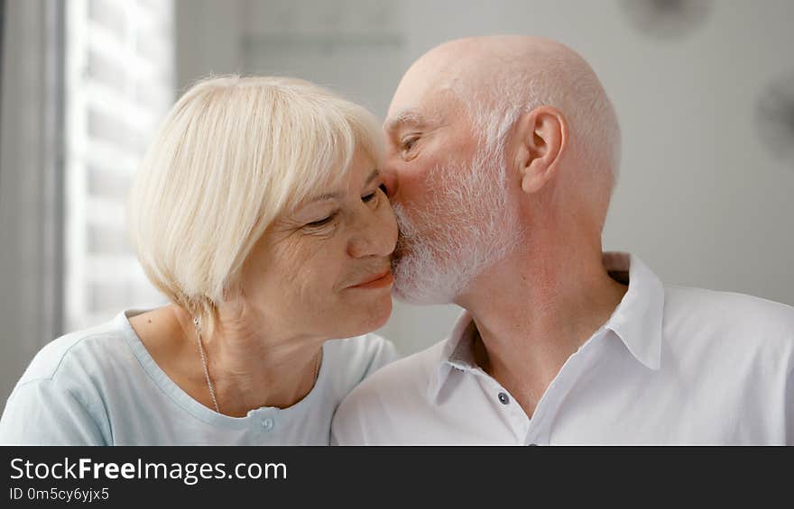 Portrait of happy senior couple at home. Senior men expresses his emotions and kisses his wife. Very emotional moment. Happy family enjoying time together. Portrait of happy senior couple at home. Senior men expresses his emotions and kisses his wife. Very emotional moment. Happy family enjoying time together.