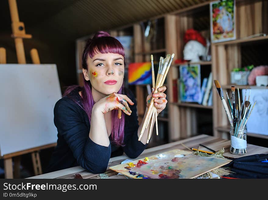 Beautiful and serious female artist with purple hair and dirty hands with different paints on them, holding paintbrushes in her art studio.