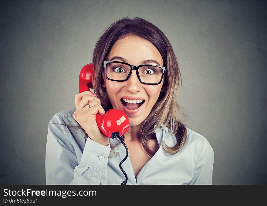 Portrait of a happy amazed woman talking on the red phone over gray wall background