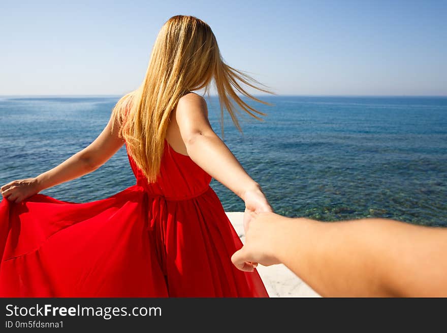 Follow me. Young blond woman in the red dress holding the friend`s hand at the beach in Cyprus.