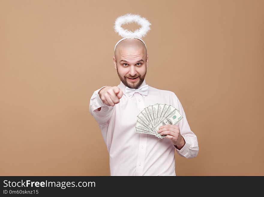 This is for you. Happy handsome middle aged bald bearded angel in shirt and white halo on head holding many dollars, pointing and looking at camera. studio shot, isolated on light brown background.
