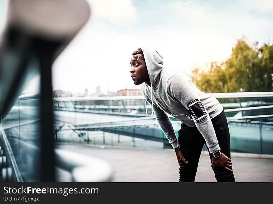 Young sporty black man runner with smartwatch, smartphone and earphones on the bridge in a city, resting. Young sporty black man runner with smartwatch, smartphone and earphones on the bridge in a city, resting.