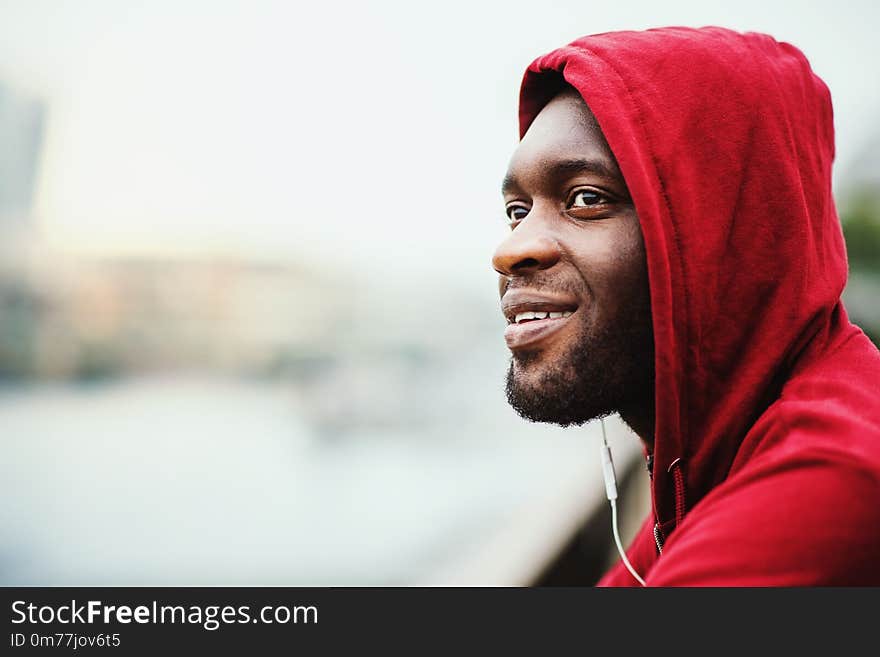 A close-up of black man runner with earphones and hood on his head standing in a city. Copy space.