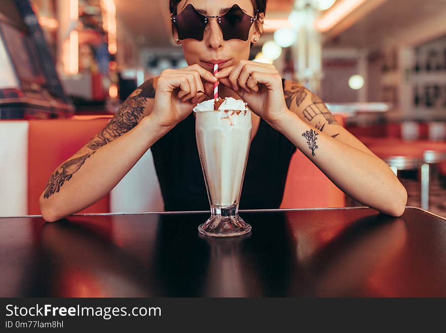 Woman wearing star shaped eyeglasses drinking milkshake at a restaurant. Woman with tattooed arms drinking milkshake with a straw at a diner. Woman wearing star shaped eyeglasses drinking milkshake at a restaurant. Woman with tattooed arms drinking milkshake with a straw at a diner.