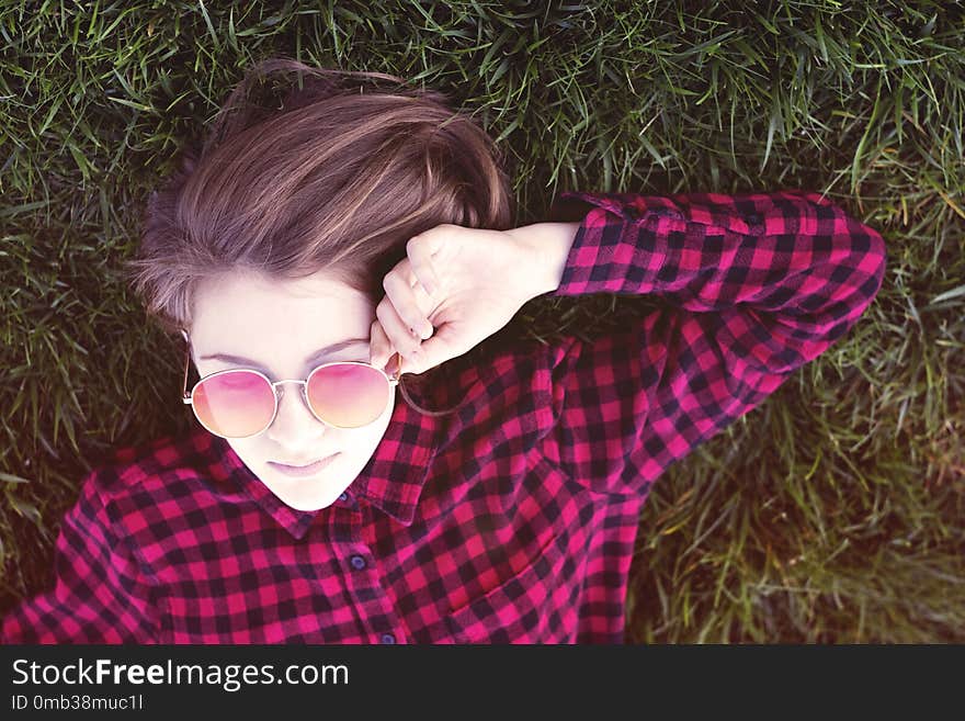 Sweet teenage girl laying on grass in park in trendy red glasses and posing for camera. Casual young woman on green grass. Sweet teenage girl laying on grass in park in trendy red glasses and posing for camera. Casual young woman on green grass.