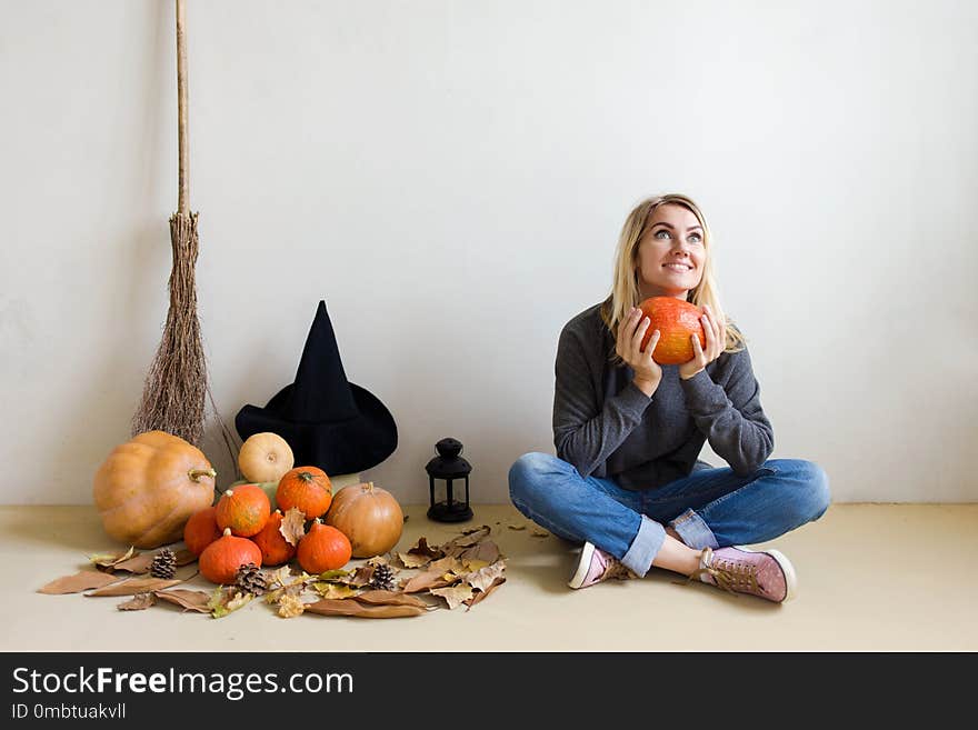 Halloween concept. Beautiful blonde girl with a witch hat with pumpkins and a broom sitting on the floor, holding a pumpkin and smiling. Against a white wall Copy space. Halloween concept. Beautiful blonde girl with a witch hat with pumpkins and a broom sitting on the floor, holding a pumpkin and smiling. Against a white wall Copy space