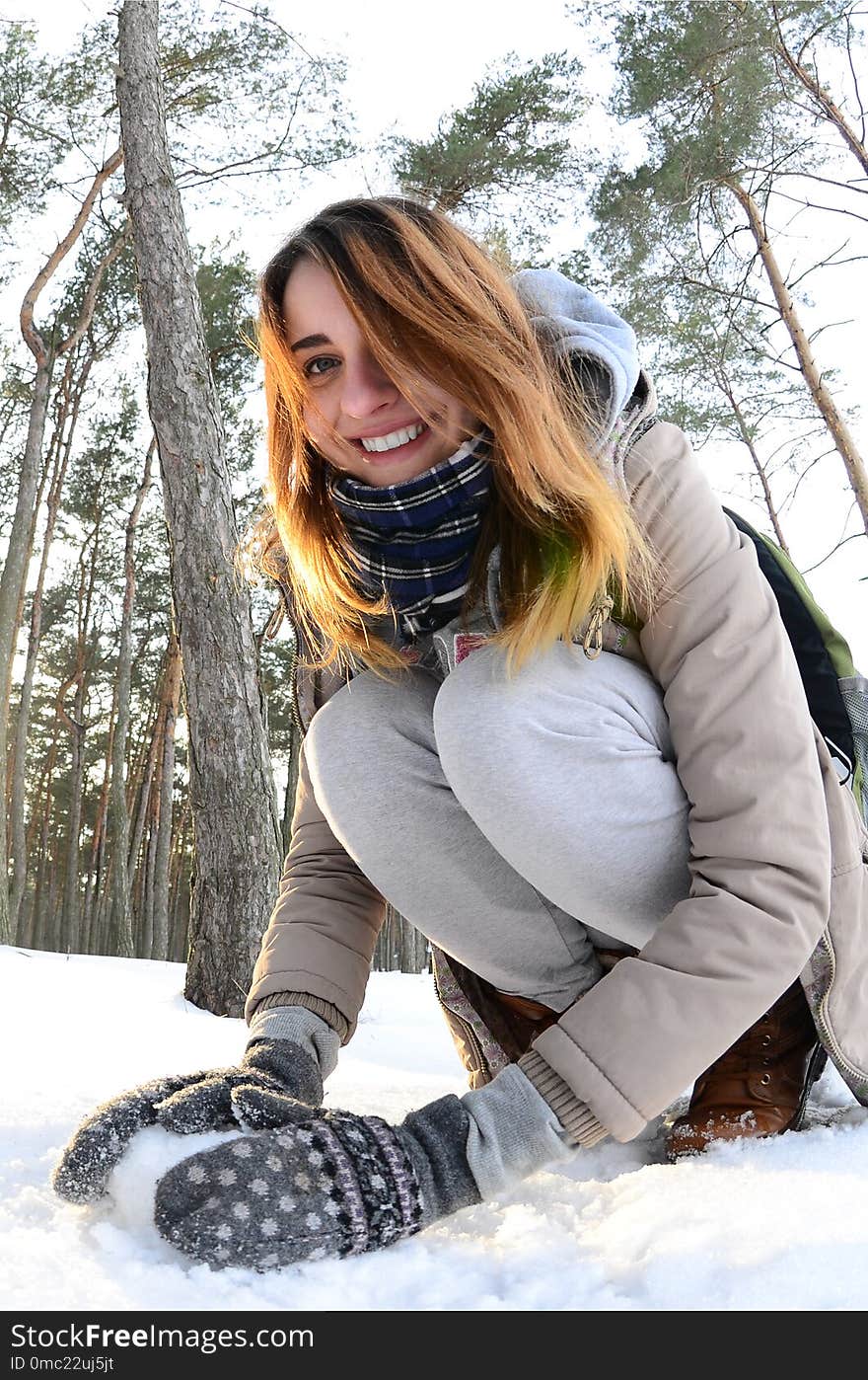 A young and joyful Caucasian girl in a brown coat sculpts a snowball in a snow-covered forest in winter. Games with snow in the open air. Fisheye Photo