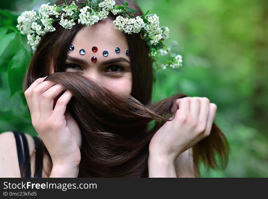 A forest picture of a beautiful young brunette of European appearance with dark brown eyes and large lips. On the girl`s head is wearing a floral wreath, on her forehead shiny decorations