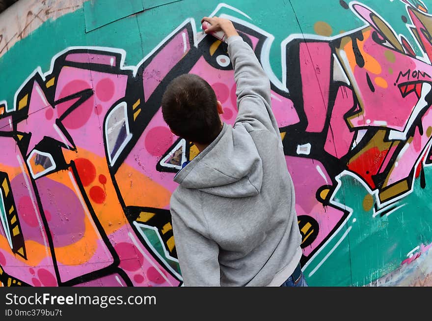 A young guy in a gray hoodie paints graffiti in pink and green colors on a wall in rainy weather. Fisheye shot