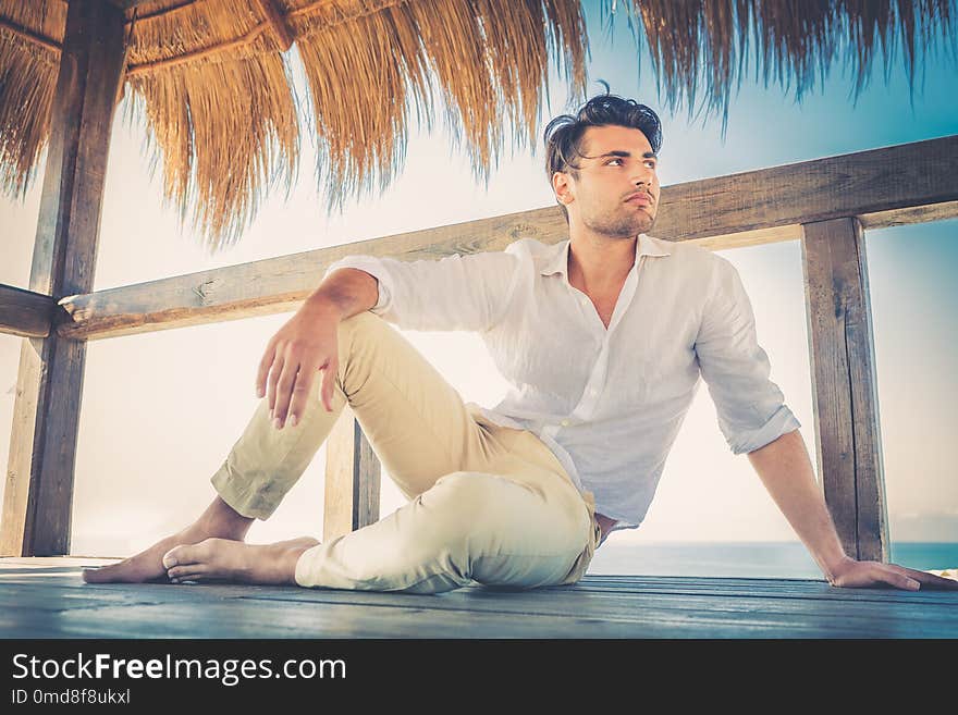 Beautiful young relaxed man in a small wooden deck. Strong summer warm lights. Trendy haircut. White shirt and beige trousers.