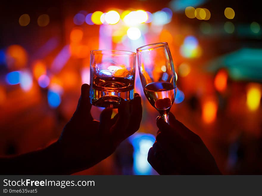 Wineglass Of Champagne In Woman Hand And A Glass Of Whiskey In A Man Hand Against A Background Of Colored Lights