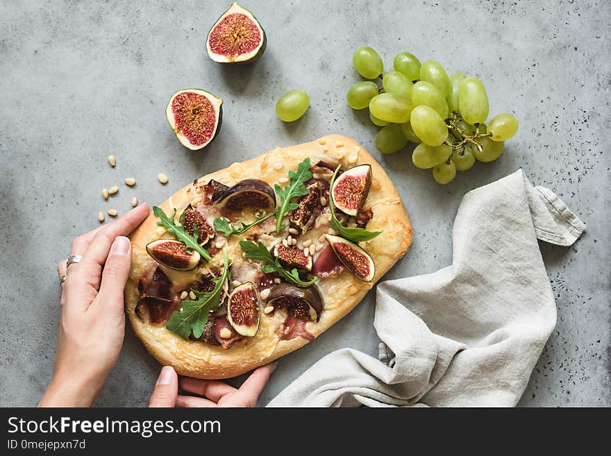 Flatbread pizza with figs, arugula and prosciutto. Hands holding fresh homemade pizza over concrete background, top view