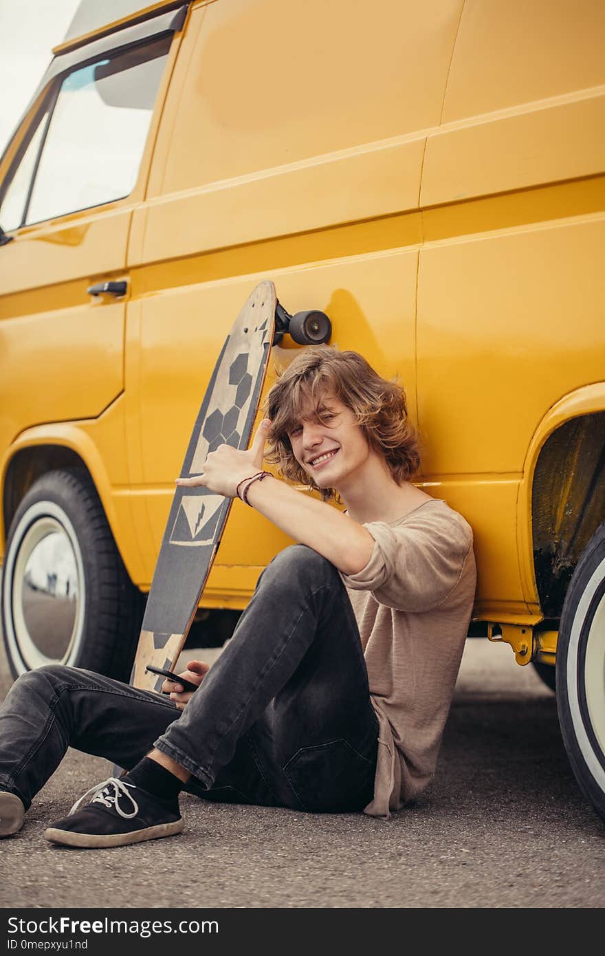 Man sitting on asphalt near his travel car with longboard, preparing to trip. Man sitting on asphalt near his travel car with longboard, preparing to trip