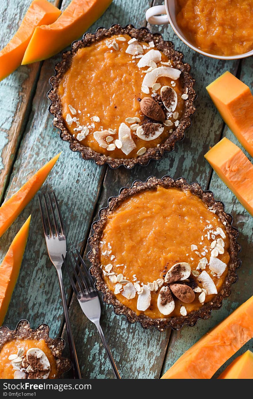 Healthy vegan dessert. Homemade tarts with nuts and oatmeal crust and pumpking filling served with raw pumpkin on blue background above view