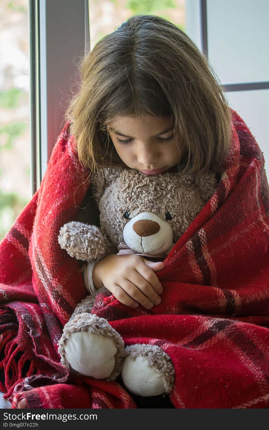 Sad little girl sitting on the windowsill hiding behind an old checkered plaid and carefully embracing her beloved toy bear. Sad little girl sitting on the windowsill hiding behind an old checkered plaid and carefully embracing her beloved toy bear