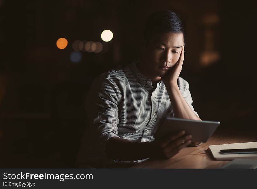 Tired young Asian businessman using a digital tablet while sitting in an office late at night with city lights glowing in the background. Tired young Asian businessman using a digital tablet while sitting in an office late at night with city lights glowing in the background