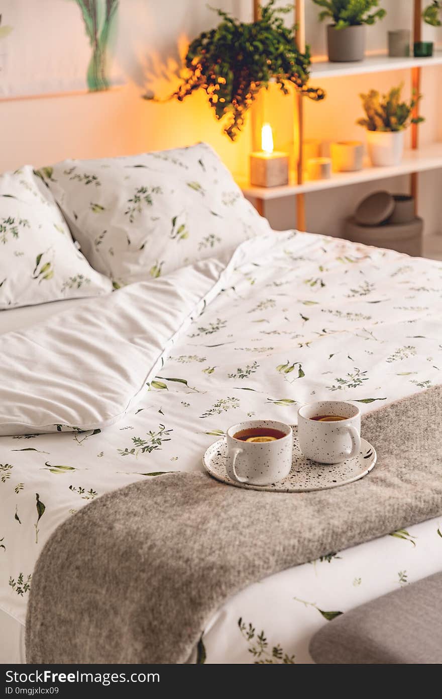 A mood created by a night lamp standing beside a bed dressed in green plants on white pattern linen and pillows in a natural style bedroom interior. Cups of tee served on the bed. Real photo