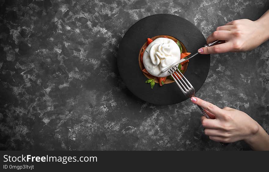 Woman eating burrata cheese on small wooden plate served with fresh tomatoes and basil on dark textured background