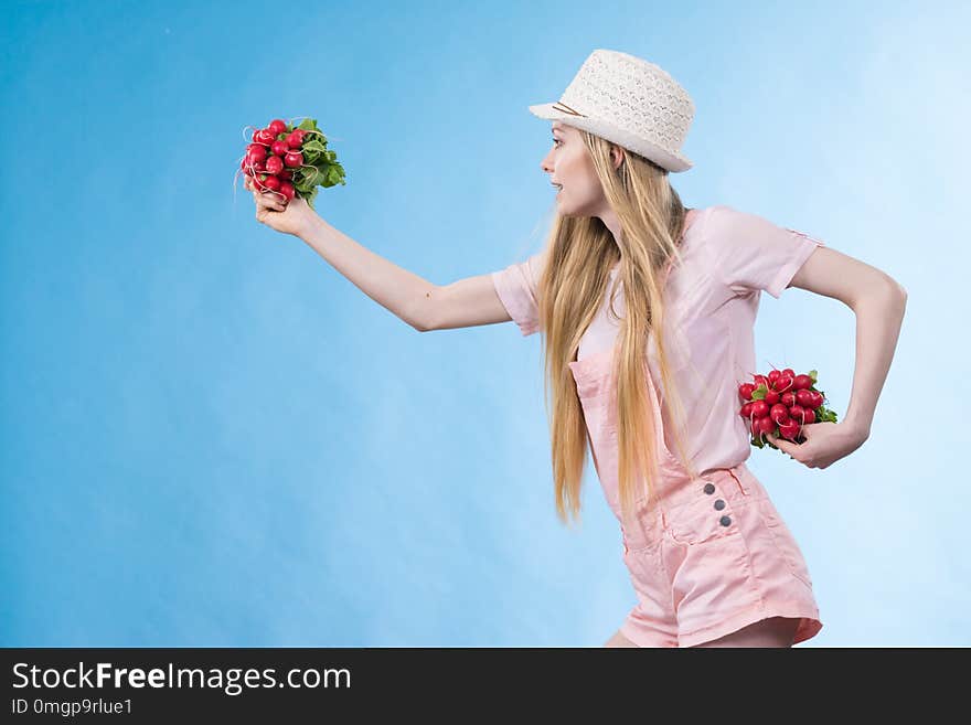 Positive teenage blonde long hair girl wearing summer clothing sun hat holding two bunches of fresh radish, on blue. Positive teenage blonde long hair girl wearing summer clothing sun hat holding two bunches of fresh radish, on blue