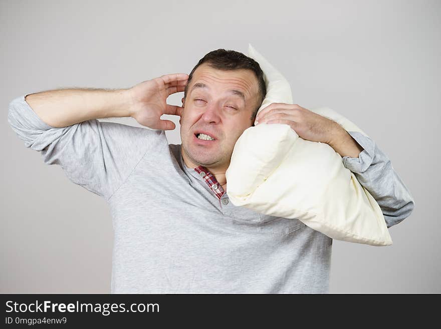 Sleepy tired angry guy holding pillow in one and closing his ear with another hand protecting from noise, on grey. Health care, sleep disorder concept. Sleepy tired angry guy holding pillow in one and closing his ear with another hand protecting from noise, on grey. Health care, sleep disorder concept