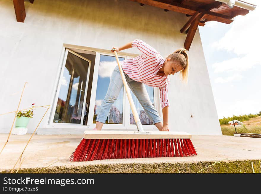 Female adult young woman using big broom to clean up backyard patio. Housecare duties concept. Female adult young woman using big broom to clean up backyard patio. Housecare duties concept.