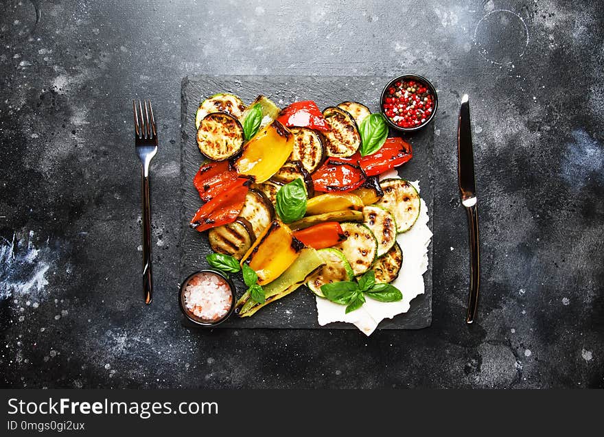 Grilled multicolored vegetables, aubergines, zucchini, pepper with green basil on serving stone board on gray background, top view