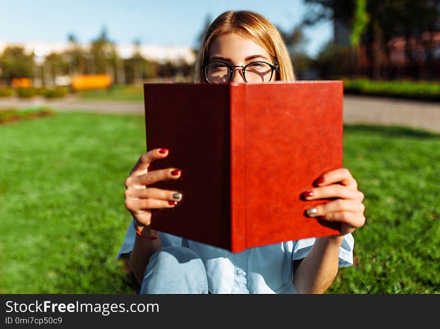 Portrait of cheerful student girl sitting outdoors on green grass, wearing glasses, holding book near her face. Portrait of cheerful student girl sitting outdoors on green grass, wearing glasses, holding book near her face