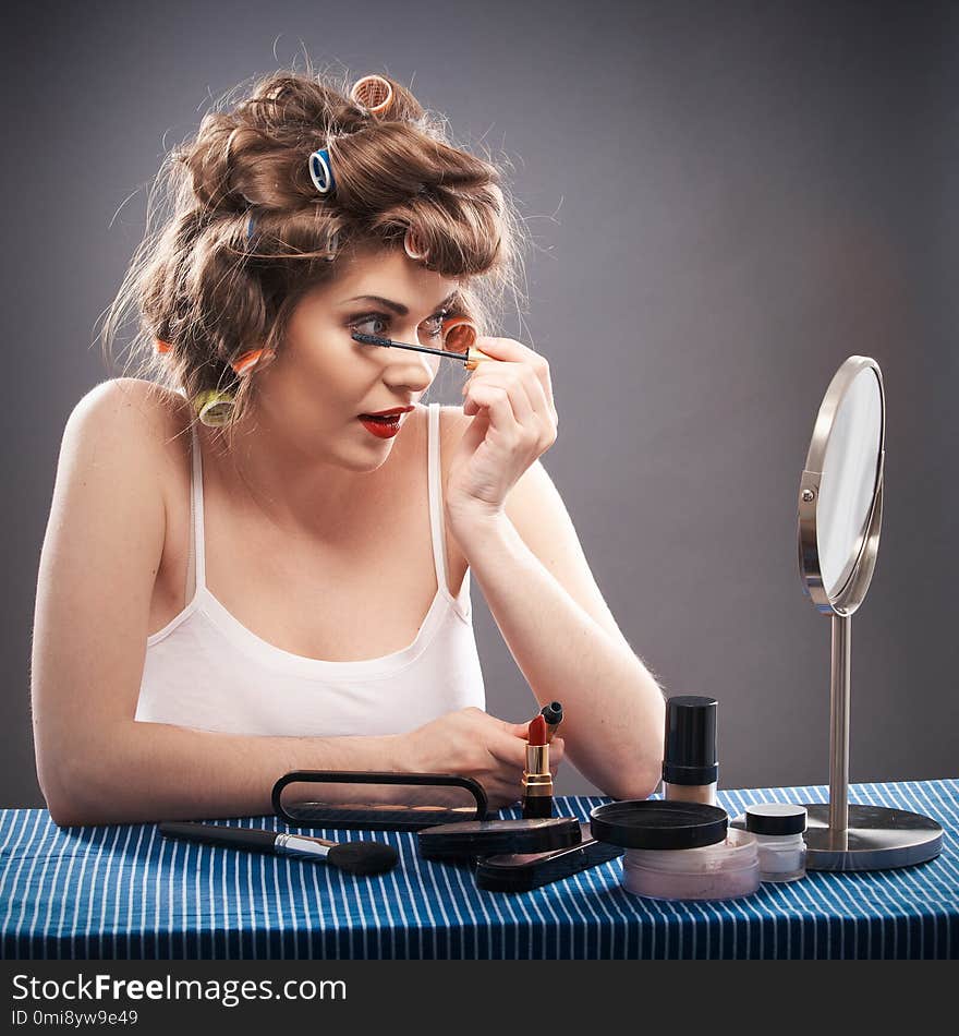 Portrait of a young woman with long hair on gray background making beauty face and hair style. Smile happy girl seating at table with make up accessories and mirror, creating home beauty salon. Portrait of a young woman with long hair on gray background making beauty face and hair style. Smile happy girl seating at table with make up accessories and mirror, creating home beauty salon