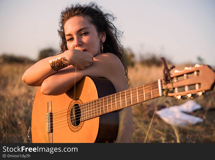 Naked Contemplative Curly brunette girl Sitting with guitar in the field with taked off clothes on background