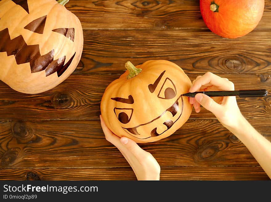 Woman`s hands drawing scary and funny Jack O Lantern face on ripe orange pumpkin with black marker pen for all hallow eve Halloween party. Holiday decoration concept. Close up, copy space, background. Woman`s hands drawing scary and funny Jack O Lantern face on ripe orange pumpkin with black marker pen for all hallow eve Halloween party. Holiday decoration concept. Close up, copy space, background.