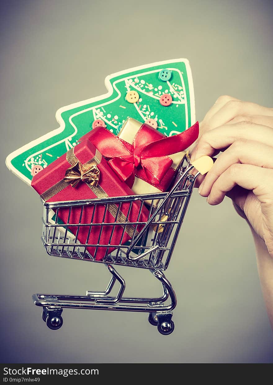 Xmas, seasonal sales, winter celebration concept. Woman hand holding shopping trolley cart basket with little christmas tree and gifts inside. Xmas, seasonal sales, winter celebration concept. Woman hand holding shopping trolley cart basket with little christmas tree and gifts inside.