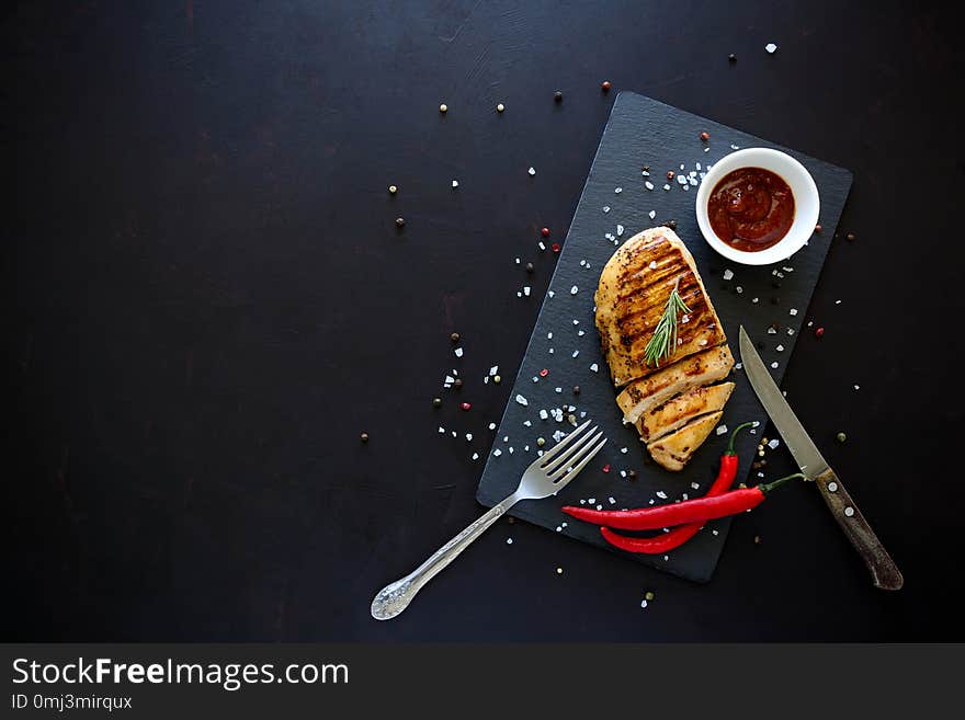 Grilled chicken fillets on slate plate with rosemary, pepper, ketchup and spices on dark wooden background. Top view. Flat lay.