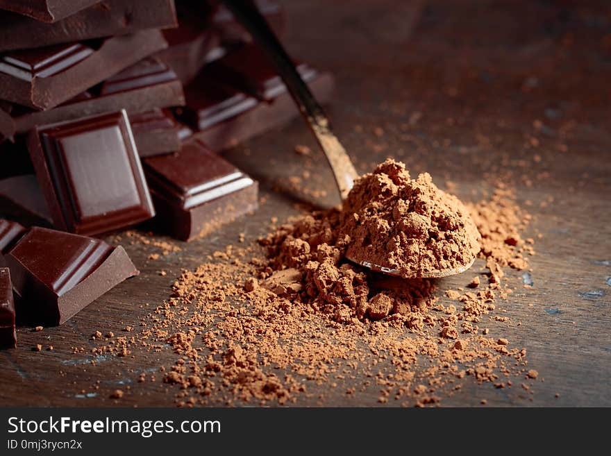 Broken chocolate pieces and cocoa powder in small spoon on a wooden background. Copy space. Broken chocolate pieces and cocoa powder in small spoon on a wooden background. Copy space.