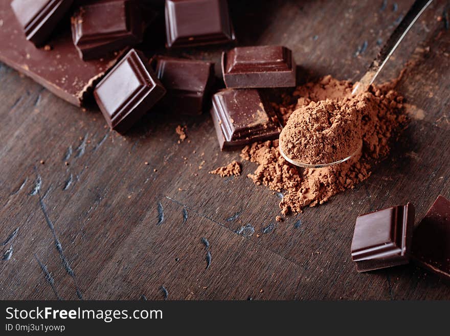 Broken chocolate pieces and cocoa powder in small spoon on a wooden background. Copy space. Broken chocolate pieces and cocoa powder in small spoon on a wooden background. Copy space.