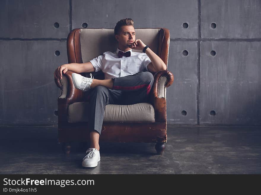 Young attractive serious glamorous male hipster in white shirt and butterfly sitting in chair, on gray background