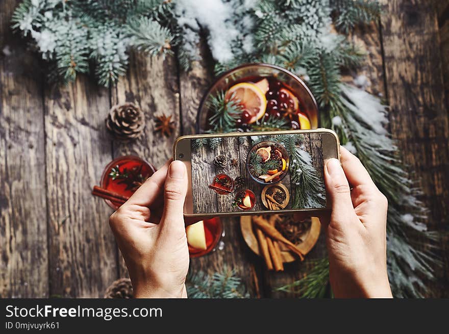 Woman taking pictures of of glass bowl with Christmas mulled wine and ingredients on rustic wooden background with spruce branches using phone. Woman Food photographer with hands visible photographing for the social networks. Woman taking pictures of of glass bowl with Christmas mulled wine and ingredients on rustic wooden background with spruce branches using phone. Woman Food photographer with hands visible photographing for the social networks.