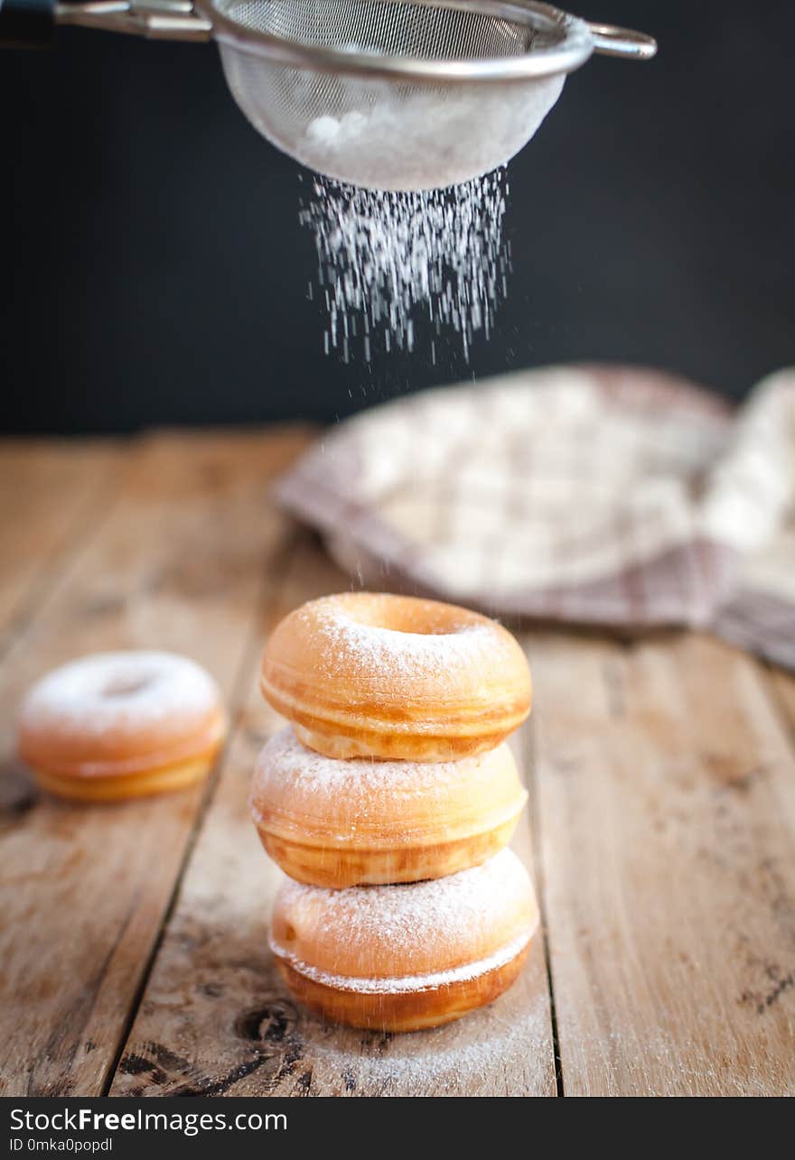 Sprinkling powdered sugar from above on stacked homemade donuts, on wooden background. Sprinkling powdered sugar from above on stacked homemade donuts, on wooden background.