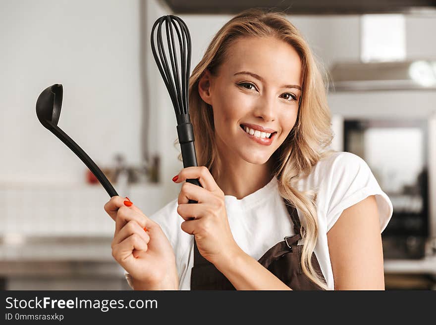 Smiling young woman chef cook in apron standing at the kitchen, holding ladle and whisk