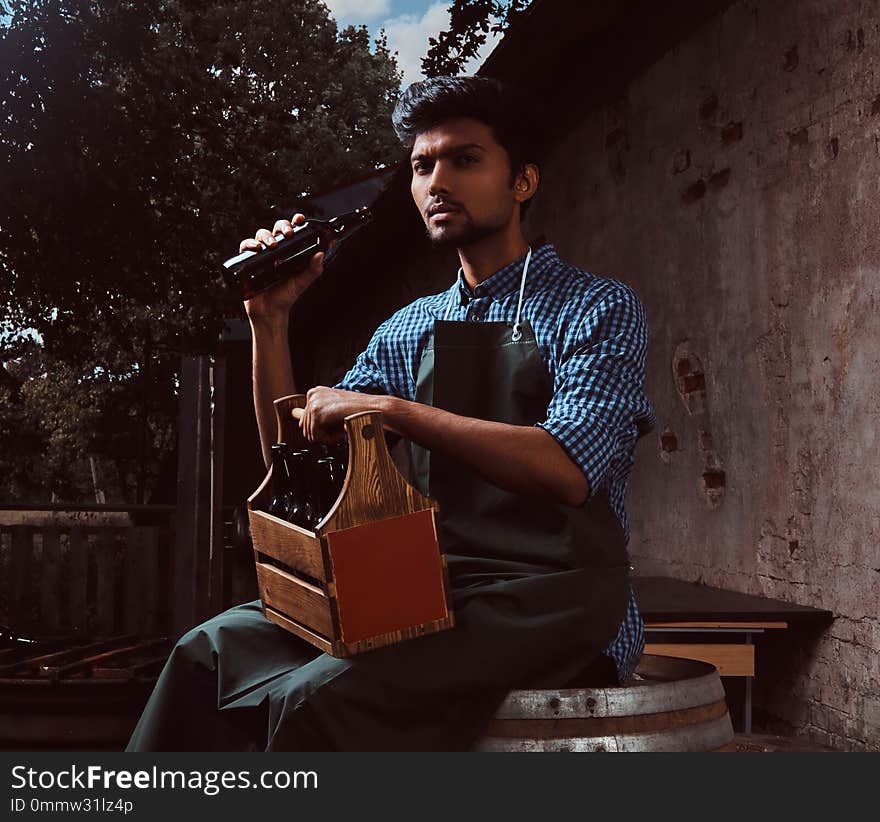 Indian brewer in uniform sitting on a wooden barrel and drink craft beer.