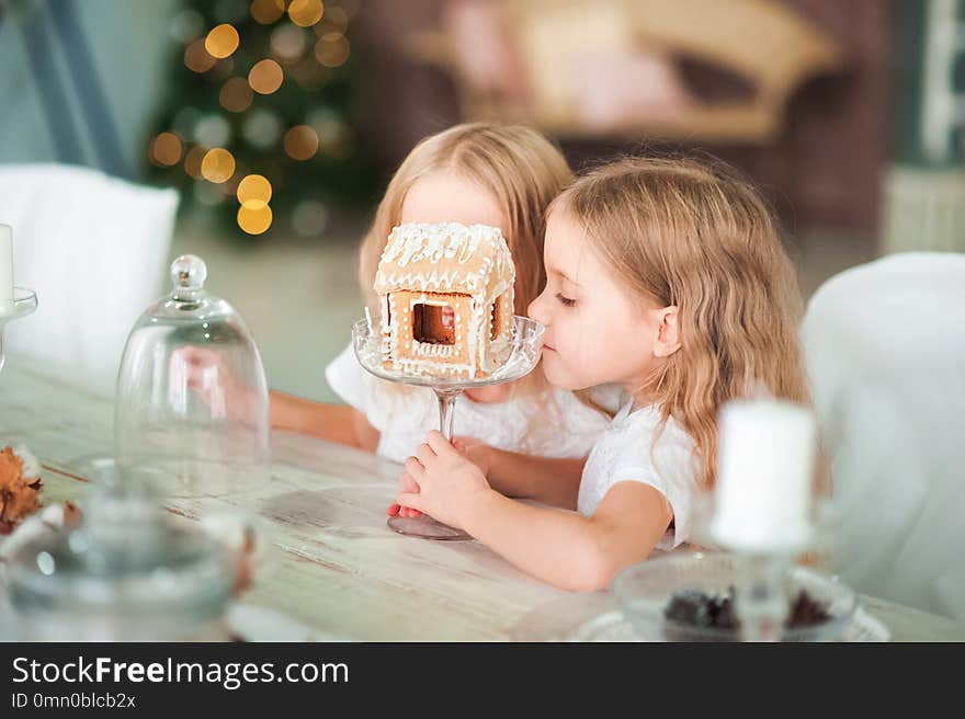 Two twin sisters at a table in the kitchen playing with a ginger house for the New Year. The kitchen in bright colors is decorated for Christmas