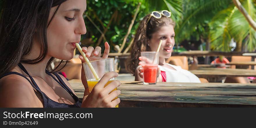 Two girlfriends sitting in bar cafe, drinking fruit cocktail on a tropical beach. Two girlfriends sitting in bar cafe, drinking fruit cocktail on a tropical beach