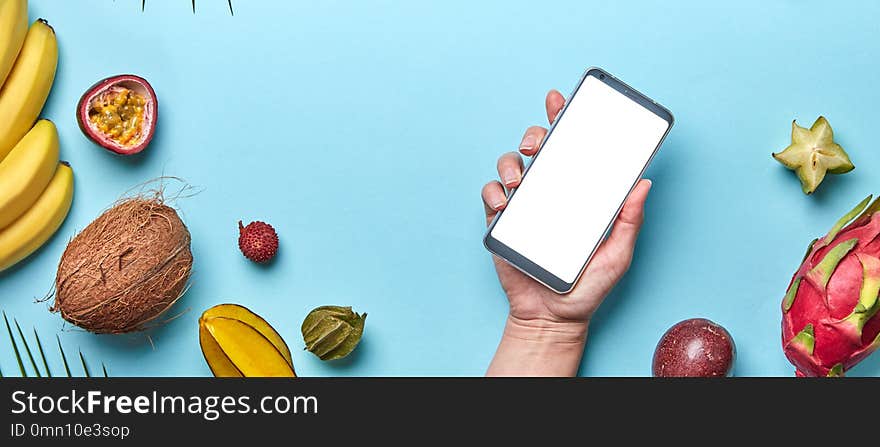 A variety of tropical fruits and a female hand with a phone on a blue background with copy space. The concept of buying products on the Internet. Flat lay. A variety of tropical fruits and a female hand with a phone on a blue background with copy space. The concept of buying products on the Internet. Flat lay