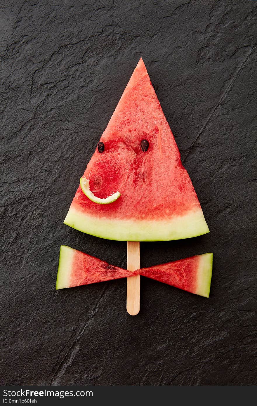 A piece of watermelon on a stick. A smiling face made with a piece of watermelon on a dark slate background with space for text. Flat lay. A piece of watermelon on a stick. A smiling face made with a piece of watermelon on a dark slate background with space for text. Flat lay